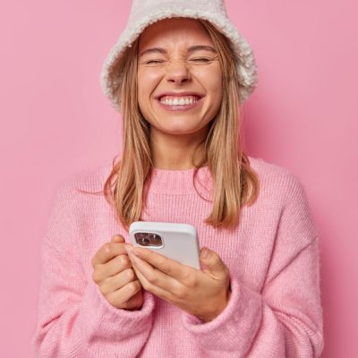 woman-squints-smiles-gladfully-glad-receieve-message-wears-casual-jumper-hat-satisfied-with-amazing-application-pink